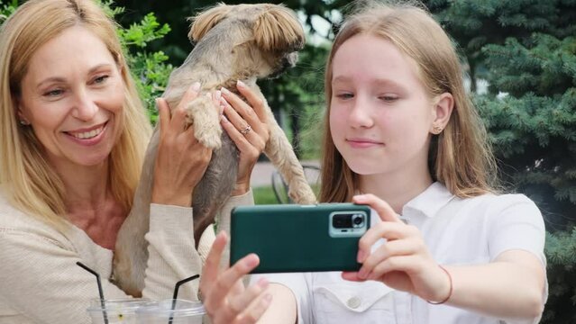 Happy mother and daughter make selfie with Shih tzu dog in park cafe. Cheerful family with pet takes picture on restaurant terrace