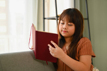 Happy Asian girl n casual clothes reading a book on couch at home.