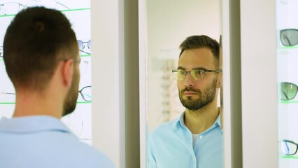 Happy young male client wearing new glasses, looking in the mirror standing near rack and showcase with eyewear. Satisfied Customer