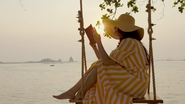 Asian woman relax on swing at the beach on bright beautiful sunset, reading book and break on the weekend with literature and peace. Female with hobby, leisure, read for knowledge and information.