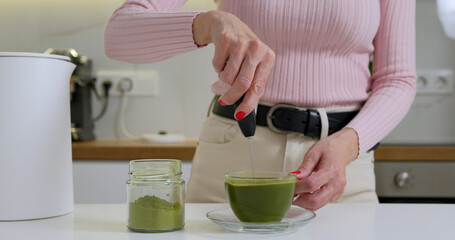 Woman is mixing matcha powder with hot water in glass cup by electric whisk 