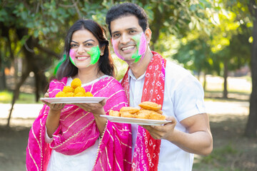 Happy mature indian couple wearing white kurta with plate of laddu and gujiyas in hands....