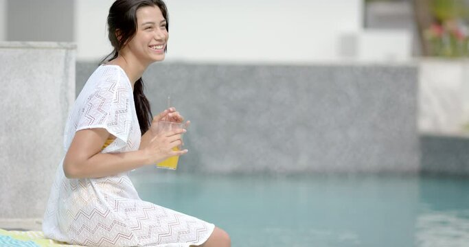 Happy biracial teenage girl holding juice sitting by pool, copy space, slow motion