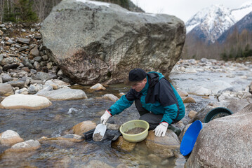 Outdoor adventures on river. Gold panning, search for gold in winter. Man is looking for gold in a mountain creek with a big boulder and blurry snow capped mountains as a background - obrazy, fototapety, plakaty