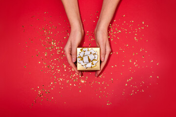 Female hands holding small gold box with a white bow gift hidden inside on red maroon background...