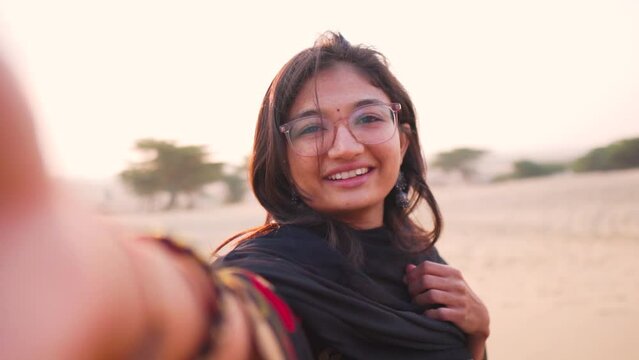 Selfie video of beautiful Indian girl standing in Thar desert wearing black Indian dress in Rajasthan, India. Tourist girl making vlog while travelling. Girl enjoying sunset time during vacation trip