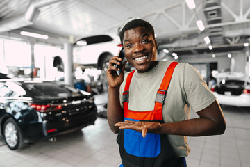 Young African mechanic man wearing uniform using mobile phone in a car service
