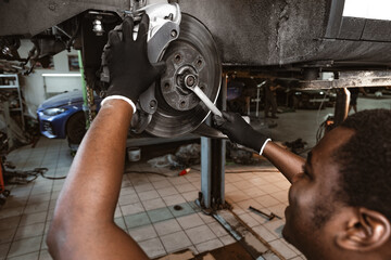 African male auto-mechanic repairing car brakes under the car in auto service - Powered by Adobe