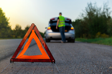 Broken silver luxury car emergency accident. Man driver installing red triangle stop sign on road. Sport automobile turned on blinkers technical problems on the road. Safety procedure when having