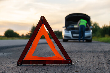 Broken silver luxury car emergency accident. Man driver installing red triangle stop sign on road....