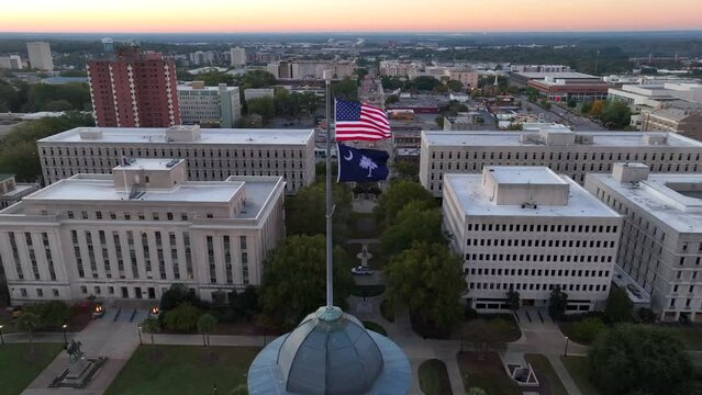 Government buildings behind American and South Carolina flags atop state house in Columbia, SC. Aerial truck shot during sunrise.