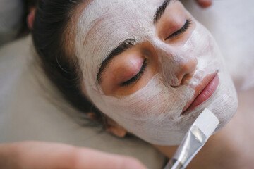 Calm relaxed young woman enjoying soothing pampering white clay facial mask for fresh clear skin. Face peeling mask, spa beauty treatment, skincare.