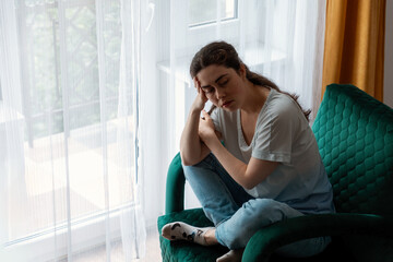 Caucasian sad young woman sitting on green velour armchair near window. Concept of depression,...
