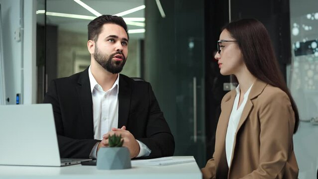 Two caucasian business partners dressed in formal clothes sitting together at office desk and talking. Charming dark haired woman attentively listening bearded man sharing ideas of new project.