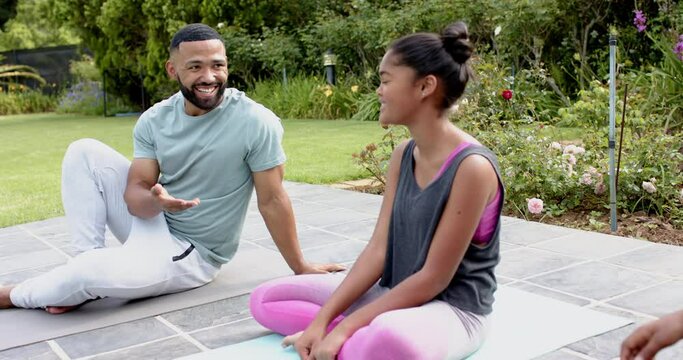 Happy african american father daughter and son doing yoga sitting in garden talking, slow motion