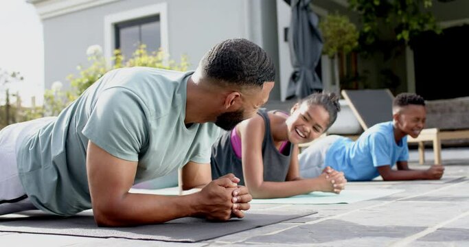 Happy african american father, son and daughter exercising in garden doing plank, slow motion