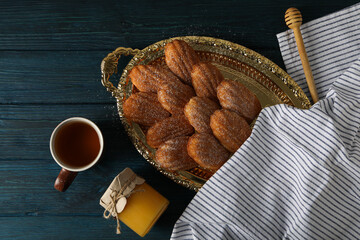 Madeleine cakes, cup of tea, jar of honey and towel on blue wooden background, top view