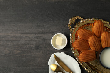 Madeleine cakes, cup of milk, cutlery and butter on wooden background, space for text