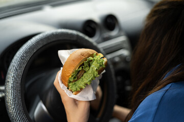 Cute woman driving car while eating hamburger on lunch. Waiting and standing in traffic jam