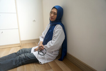 Exhausted female doctor in a gown and hijab sits on the floor of a hospital.