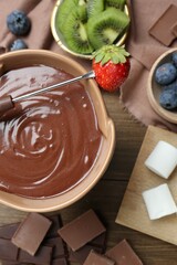 Fondue pot with melted chocolate, marshmallows, fresh kiwi, different berries and fork on wooden table, flat lay