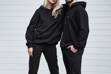 Man and woman wear black hoodies.  A fashion template for print and branding on hoodie. trendy couple on the street wearing casual apparel with no face visible. No logo sweater and pullover with hood. - obrazy, fototapety, plakaty