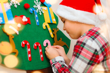 A cute seven-year-old boy in red plaid pajamas and a Santa hat decorates his felt hand-made...