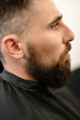 Obraz premium A young man with a beard in a black cape sits on a chair in a barbershop. The client is waiting for a haircut of the beard.