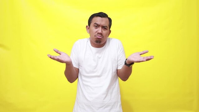 Asian man shrugs shoulders unaware raises hand clueless has no idea cannot answer poses against yellow background. Indonesian male not knowing information gesture