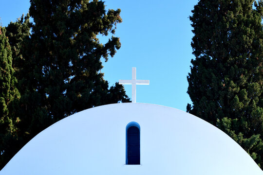 Closeup view of old white greek chapel, little church with shiny white cross in natural sunlight, blue sky and dark green cypress trees in the background, Peloponnese Peninsula, Greece, Europe