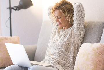 One cute adult woman using laptop sitting on the sofa in bright home. People and technology online. Modern female watching computer and relax her back and shoulders raising arms under the head - obrazy, fototapety, plakaty