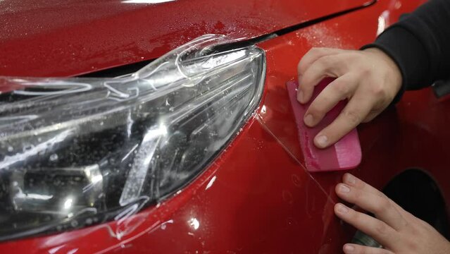 The master applies vinyl film to the headlight of a red car. Closeup view on worker detailer hand smoothing with a scraper protective film. 
