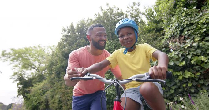 Happy african american father teaching son to ride bicycle in sunny garden, slow motion