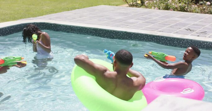 Happy african american parents, son and daughter playing with water pistols in pool, slow motion