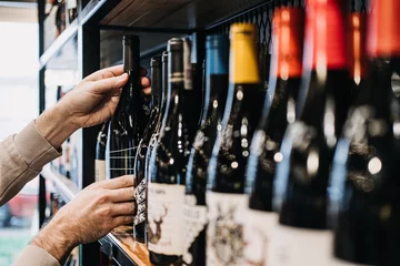 Fotobehang Customer Selecting Wine Bottle from Store Shelf. A person's hand picking a wine bottle from a diverse selection on a well-stocked wine shop shelf © irissca