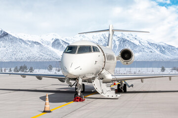 Modern white private jet plane with an opened gangway door at the winter airport apron on the background of high scenic snow capped mountains