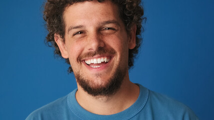 Close up, guy with curly hair dressed in blue t-shirt looks at the camera and laughs isolated on...