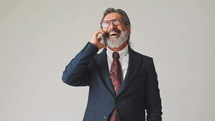 Businessman in glasses with gray hair, talking on mobile phone with a laugh, isolated on gray...
