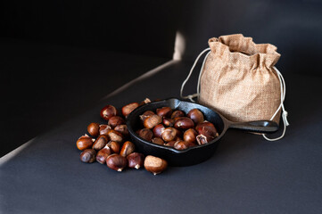 Pan with fried edible chestnuts, jute sack on black table background with sunlight ray. Traditional healthy organic autumn and winter food. Soft selective focus