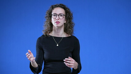 Woman speaking toward camera while standing on blue background. 20s female person looking at camera...