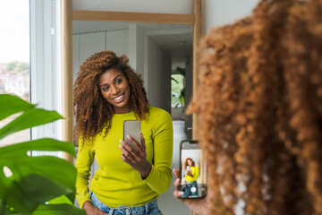 Cheerful curly woman holding mobile phone and making selfie in the mirror - 692498552