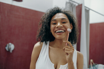 Happy woman at bathroom use facial roller for skin care and looks camera. Skin care concept