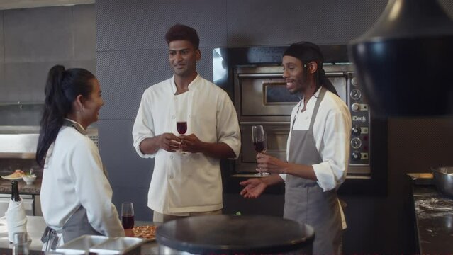Wide shot of happy sous chefs relaxing with red wine at cafe kitchen after long day at work