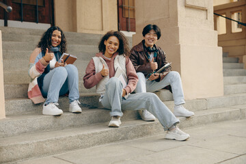 Happy students sitting on stairs outside library during study session and showing thumb up