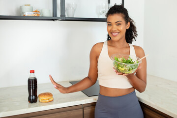 fitness lady holds salad gesturing stop to tempting burger indoors