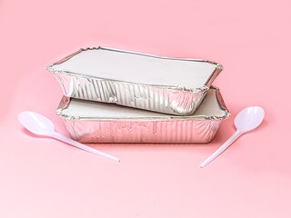 Two aluminum foil takeaway food containers with two plastic spoons on pink background. 
