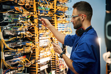 Holding smartphone. Young man is working with internet equipment and wires in server room - obrazy, fototapety, plakaty