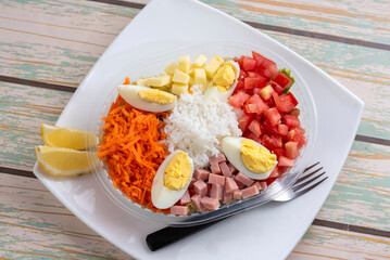 Vegetable salads with lettuce rice ham croutons tomato olives hard-boiled egg arugula carrot lemon cheese on a delivery tray on a white plate