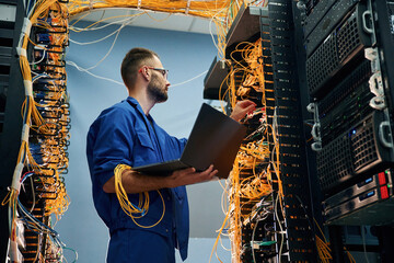 Silver colored laptop in hands. Young man is working with internet equipment and wires in server...