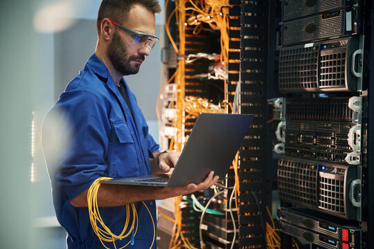Silver colored laptop in hands. Young man is working with internet equipment and wires in server room